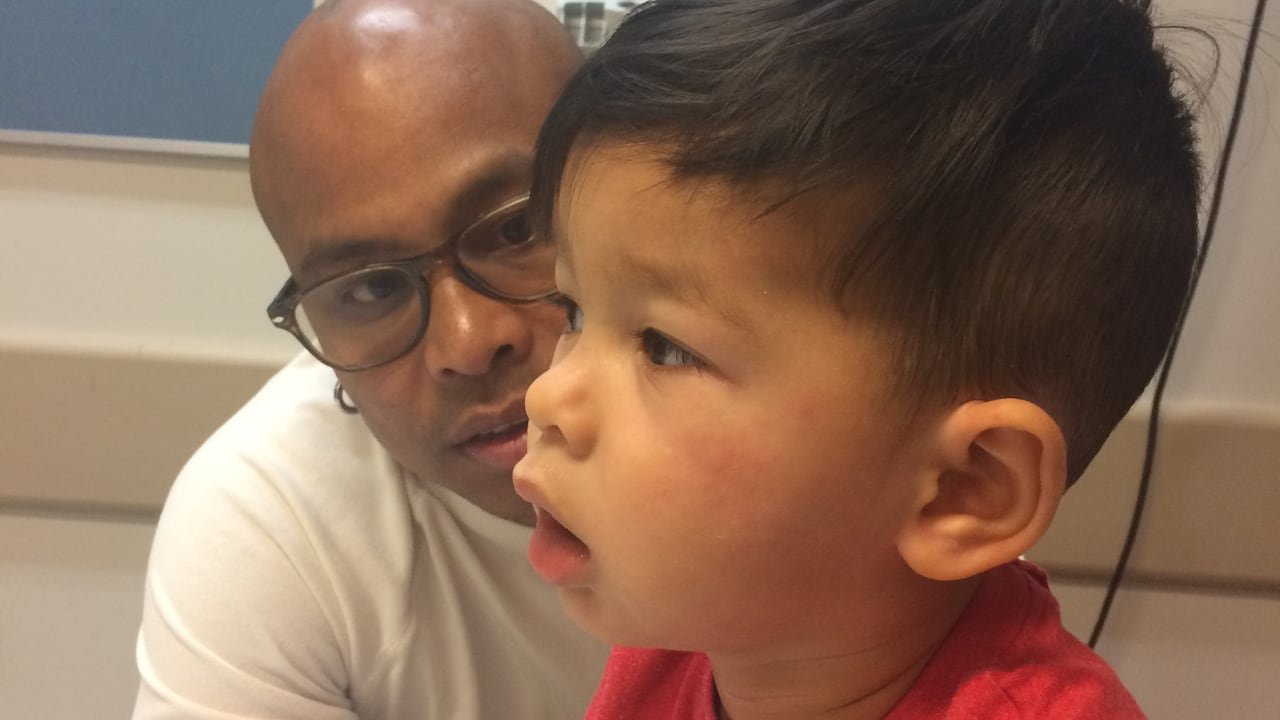 default-171 In a hospital room, a man looks concerned at a boy breathing with his mouth open and who has hives on his cheeks.