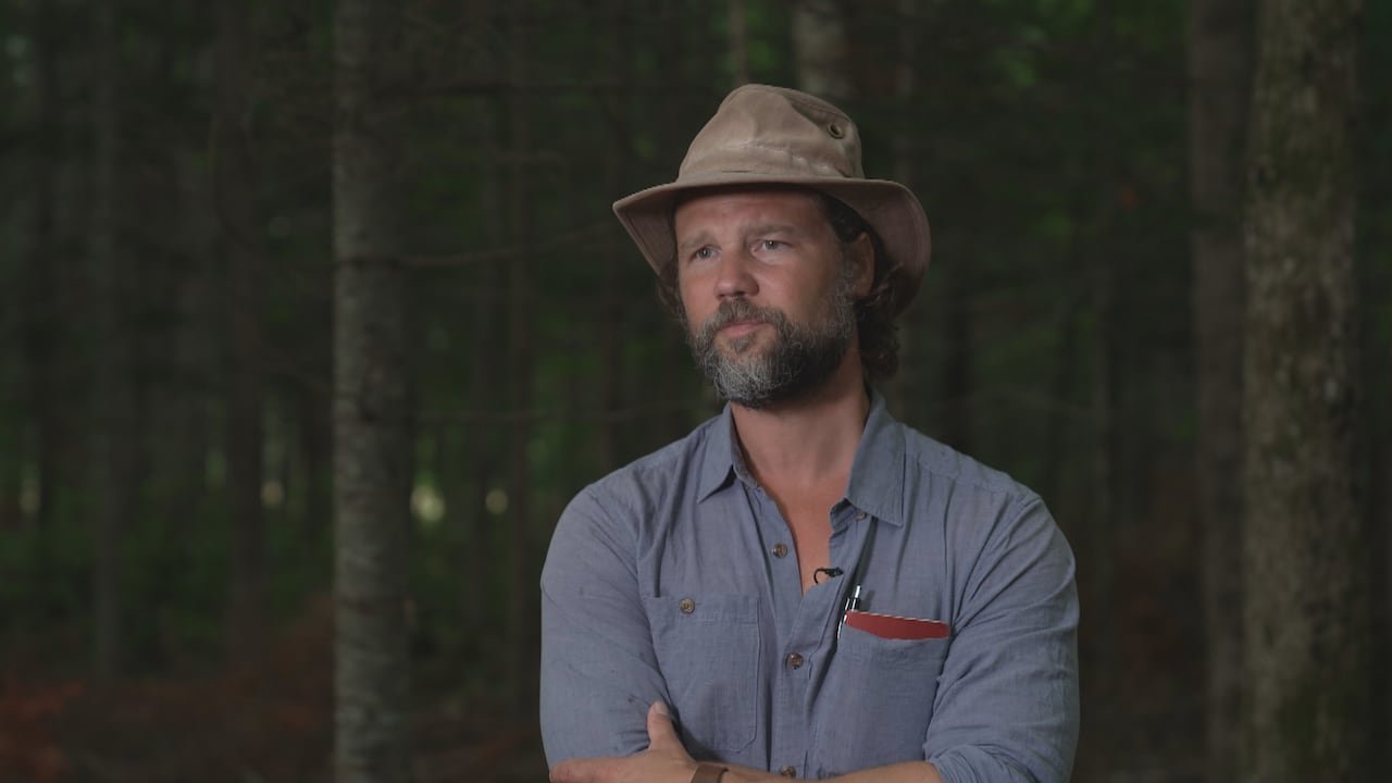 default-205 A white man with a beard, wearing a Tilley hat and a blue collared shirt, standing in front of a background of evergreen trees.
