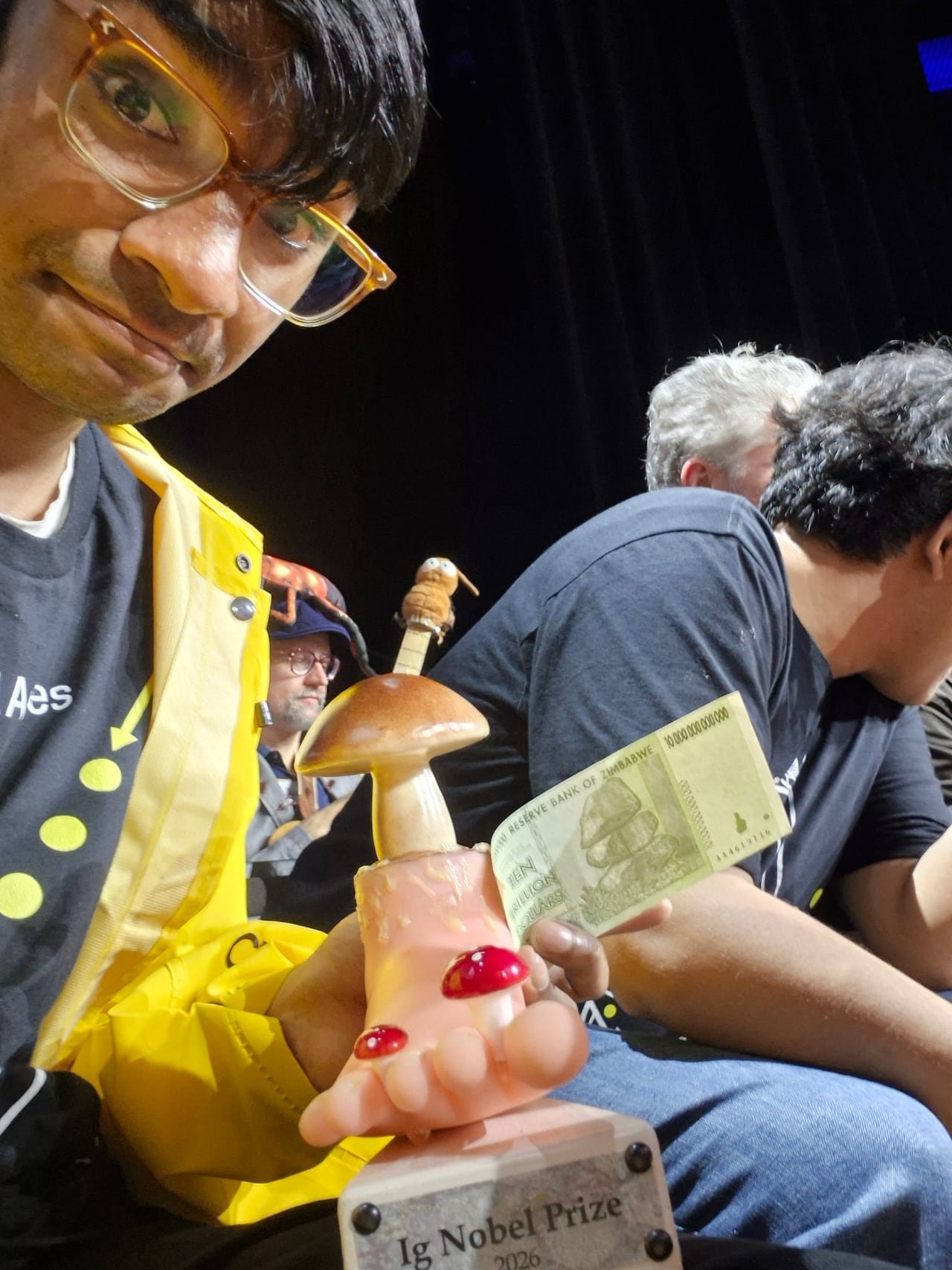 default-36 A man in glasses takes a selfie with a ten-trillion dollar bill and a trophy shaped like a foot surrounded by mushrooms.